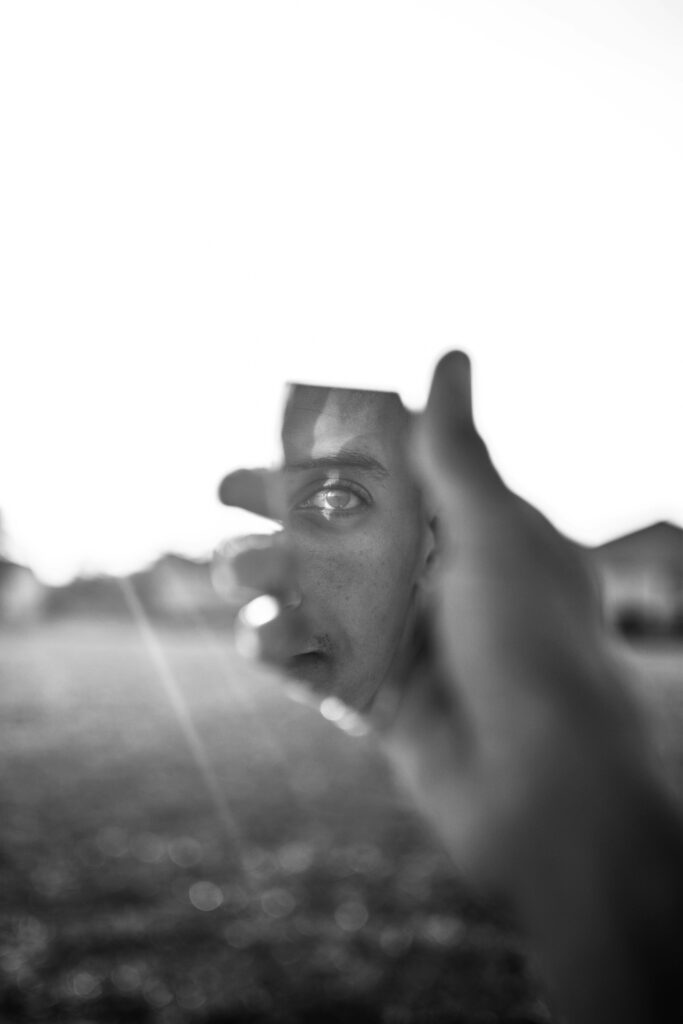 Image of a man holding a mirror shard in his hand. He stares at his reflection in the shard. In the distance, the sun shines down.