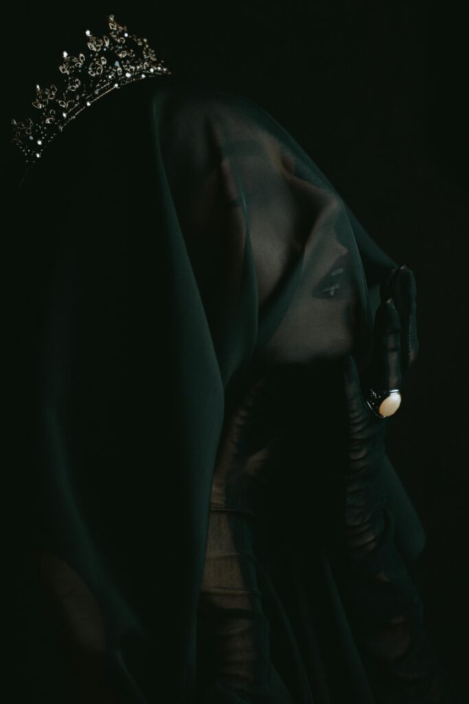 Profile of a woman whose head is covered with a black veil. Her hand, covered in a black glove, touches her face. One finger is adorned with an opal ring.