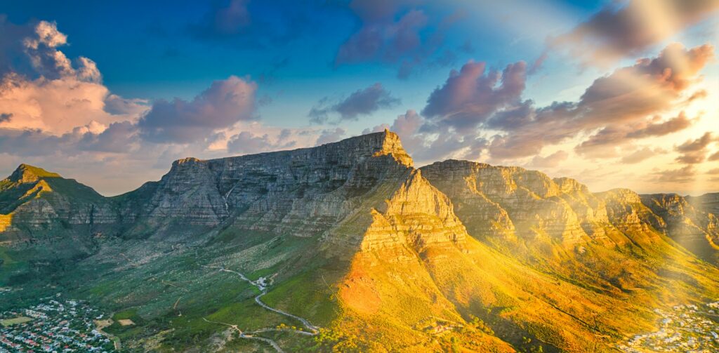 Image of the landscape of South Africa. It features massive plateaus and green valleys. Above, the sun shines across a cloudy sky.