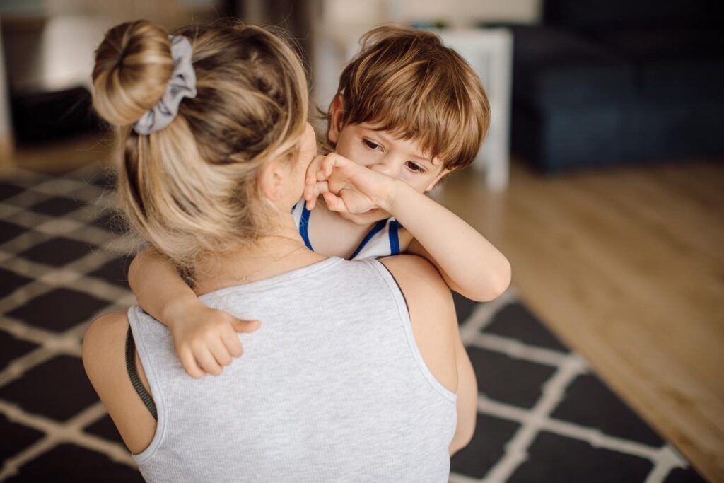 image of woman holding a small child. Her back is turned to the camera, while we see the child’s face. He is wiping his nose with the back of his hand.
