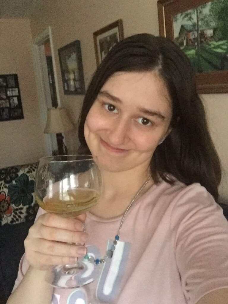 A photo of the author, Anna Bechtel, smiling and holding up a glass of white wine. 