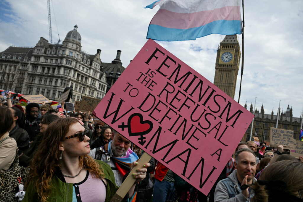 A photo of a trans rights rally on a sunny day in London, with protestors marching with signs and flags. Big Ben is visible in the background.
