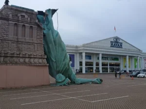 A replica of the Statue of Liberty lies toppled on its side in a parking lot outside a Havan department store. The large green statue appears broken and partially detached, resting on the ground in front of the store.
