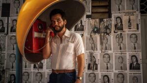 Wagner Moura stands inside a yellow phone booth, holding a red handset to his ear. He wears a light short-sleeved shirt and looks serious as he listens. Behind him, a wall is covered with repeated black and white political campaign posters featuring the same candidate’s portrait and name.
