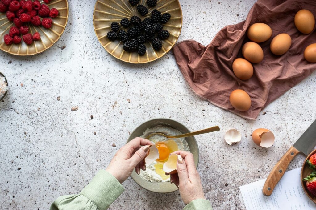 Woman cracking an egg into a bowl of sugar, surrounded by raspberries and blackberries on plates, as well as more eggs