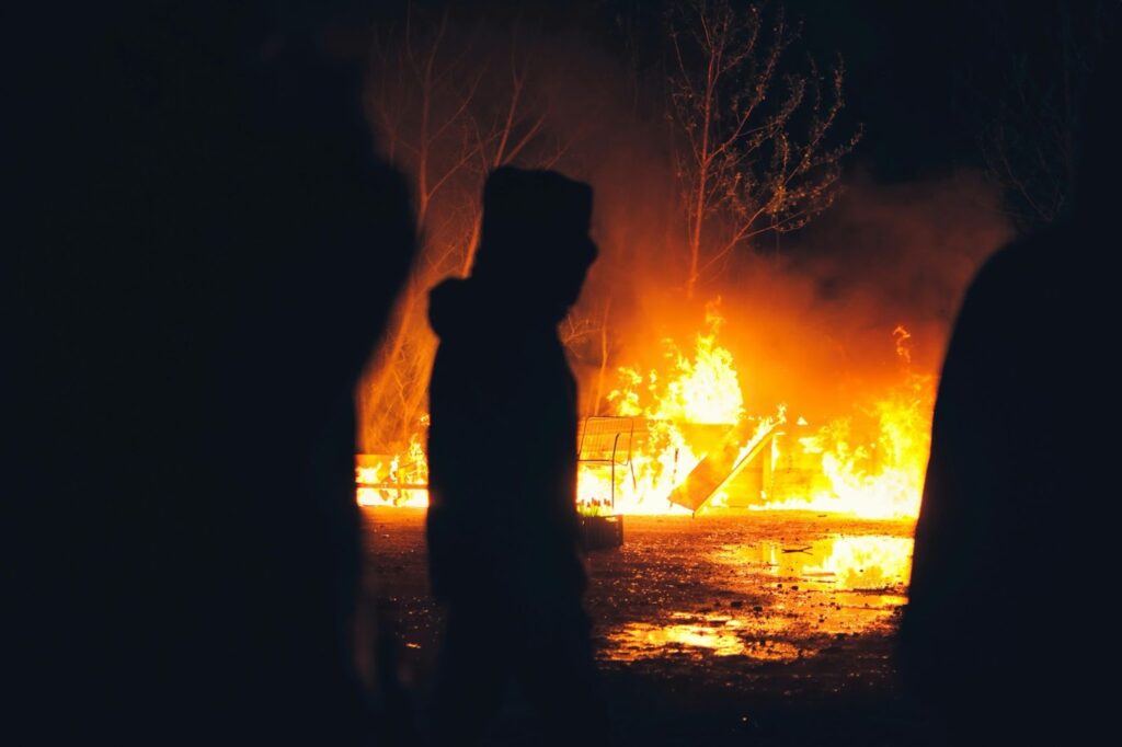 Three dark figures stand watching a public park ablaze as a bench and child’s slide go up in flames.