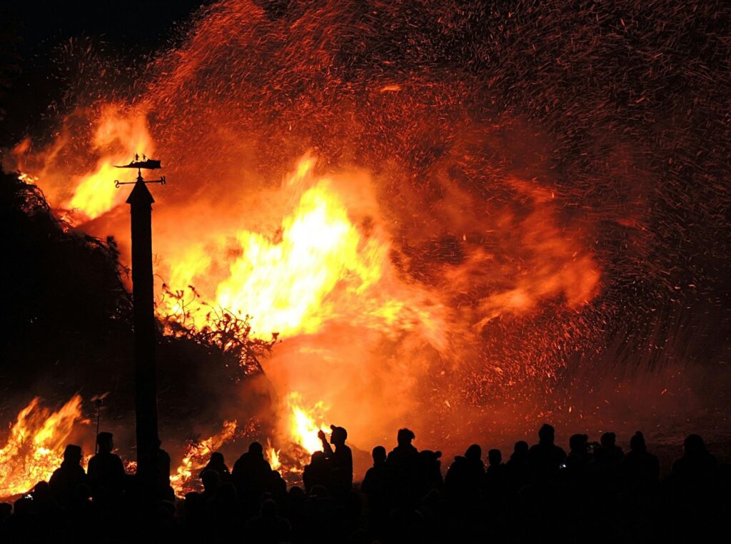 A huge crowd of people stand in the dark watching a gigantic blaze rage with sparks and flames everywhere. A small tower with a melting weathervane can be seen in contrast against the bright fire.