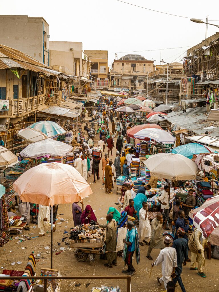 Busy outdoor Nigerian marketplace with umbrella vendors, traffic, and shoppers