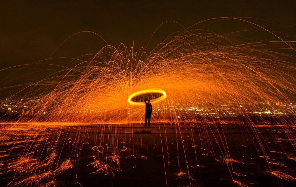 A silhouetted man stands in a swirl of flame and orange-yellow fireworks light against a dark cityscape.
