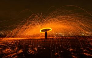 A silhouetted man stands in a swirl of flame and orange-yellow fireworks light against a dark cityscape.