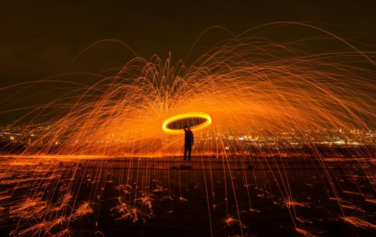 A silhouetted man stands in a swirl of flame and orange-yellow fireworks light against a dark cityscape.