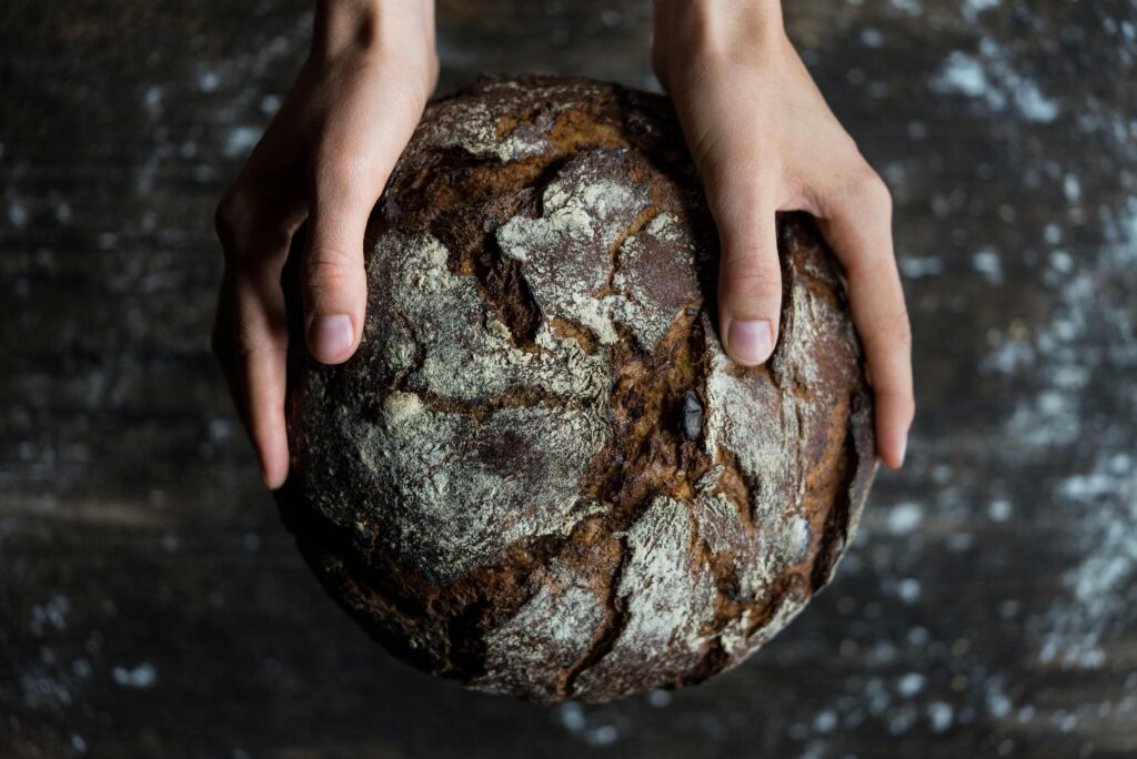 A young woman holds a dark loaf of bread. The loaf is cracked, and round like the sun.