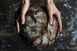 A young woman holds a dark loaf of bread. The loaf is cracked, and round like the sun.