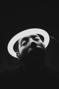 A man in shadow wears a ring light as a halo. His eyes are closed, and he looks exhausted.