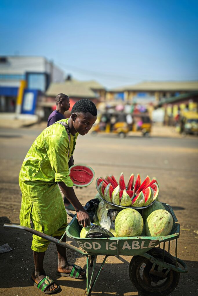 Nigerian vendor slicing watermelon into a rose in his wheelbarrow, labeled  “God is able”