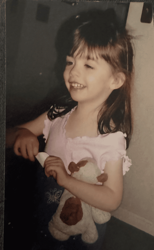 A preschool-aged girl with brown hair who is standing, holding her stuffed dog in her arms.