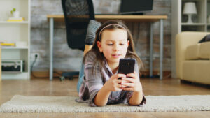 Cheerful little girl lying on the floor carpet using smartphone. | Photo by Freepik