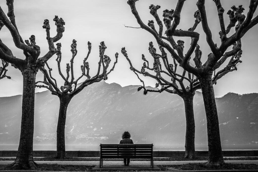 A woman sits on a bench overlooking a mountain range, surrounded by bare trees, and her stark loneliness.