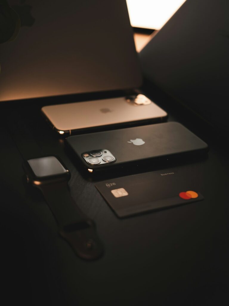 A MacBook, a smartwatch, two iPhones, and a credit card laid out on a dark table.