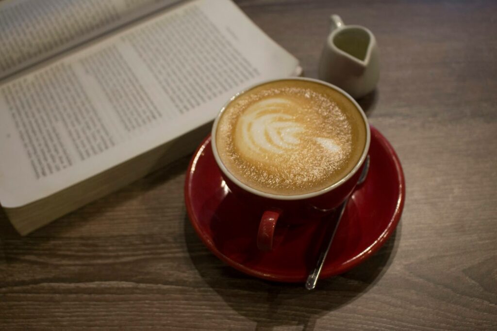 An open book on a table, next to a cup of coffee with a leaf latte art on top of a red saucer with a small creamer pitcher and spoon.