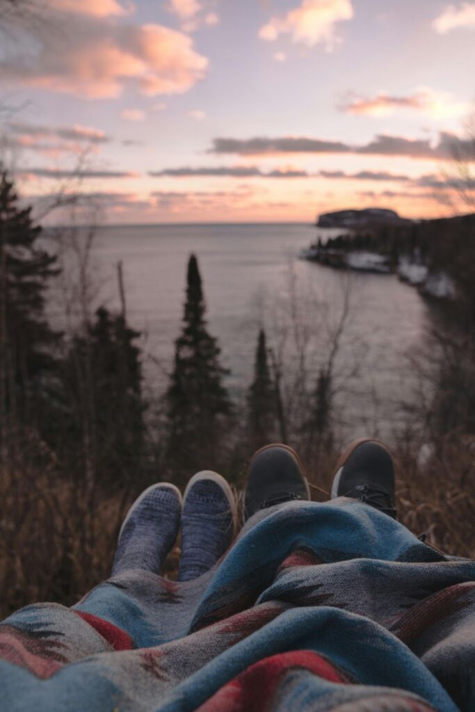 Two people sharing a blanket on a hill, enjoying the sunset over a forest and a lake.