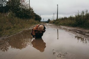 A young boy, crouched, looks at his reflection in a mud puddle. He has blond hair and is in a rust-colored jacket; the sky is gray.