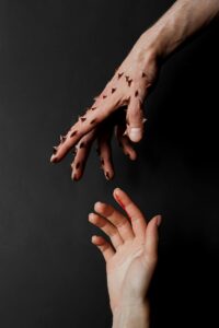 A hand, fingers outstretched, is covered in dark thorns. Another hand reaches up to grasp the first, being pricked by its thorns. Blood trickles down the second hand’s index finger.