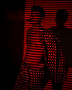 A photo of a man wearing an open striped red suit, bare-chested, standing in the shadow of blinds.