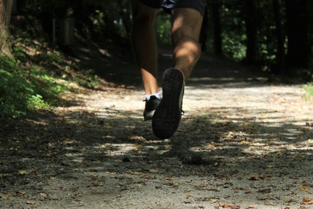 Close-up of a man’s legs and feet as he runs on a dirt path in a wooded area.