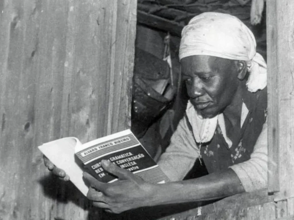 Brazilian Writer Carolina Maria de Jesus reading a book out of a small window.