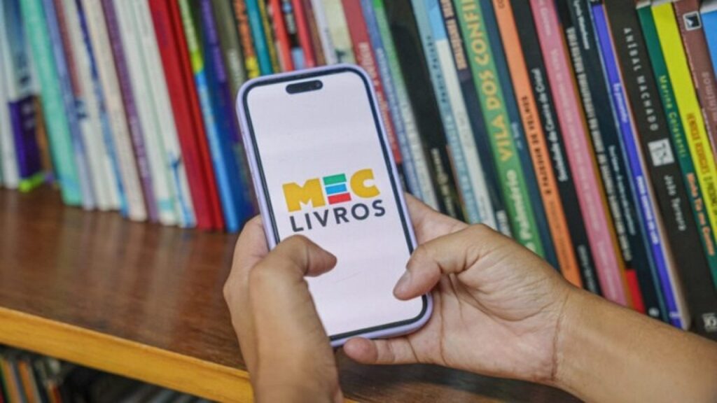 A person holding a smartphone displaying the MEC Livros platform logo in front of a bookshelf filled with books. | Photo: Brazilian Ministry of Education (MEC)/Official release