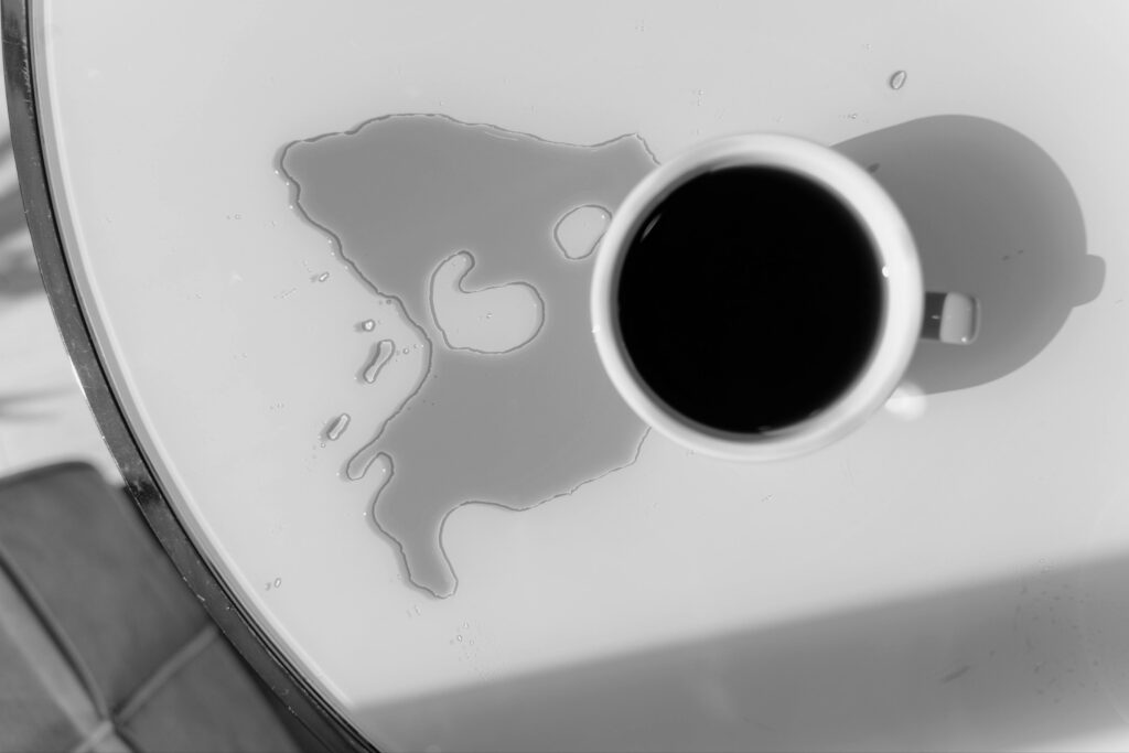 A grayscale image of a cup of coffee that has been partially spilled. The puddle of coffee on the table has gone cold.