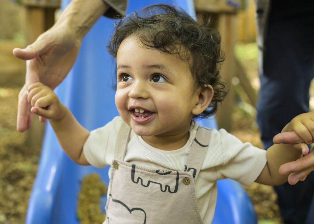 A smiling toddler holds an adult’s hands in a playground.