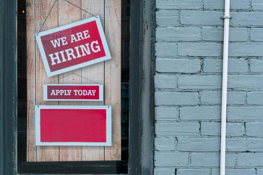 A photo of two signs hung on a wooden window of a brick wall, reading: "We are hiring,” and "Apply today."