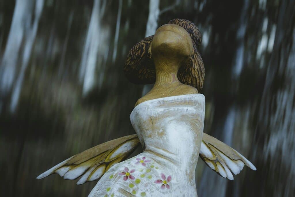 A wooden statue in a white dress with purple flowers, wings showing from behind as if in flight.
