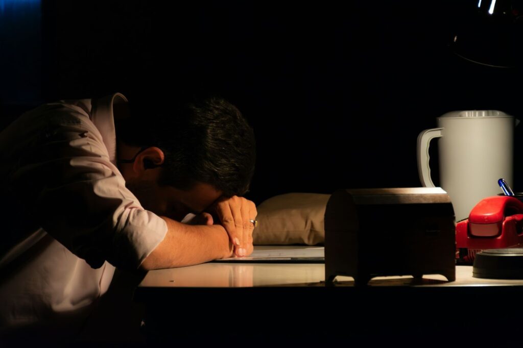 Image of a man at a table, his head down on his crossed arms in front of him. A single light illuminates his body and the objects on the table.