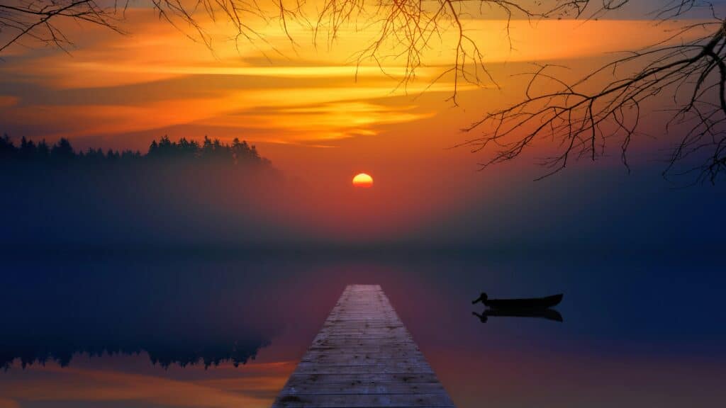 A setting sun overlooks a pier and empty boat on a foggy lake.