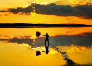 A person holds a bunch of balloons tightly in their hand at sunset. They stand in the glow of golden light, a perfect reflection of the sky, the balloons, and the person in the water.