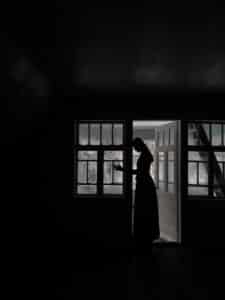 The silhouette of a woman, a mother, is shown in a dark room while winter sunlight shines through the frosted glass windows. The woman’s head rests against the doorframe, and her hand is on the windowpane.