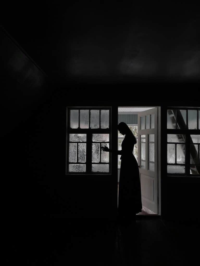 The silhouette of a woman, a mother, is shown in a dark room while winter sunlight shines through the frosted glass windows. The woman’s head rests against the doorframe, and her hand is on the windowpane.