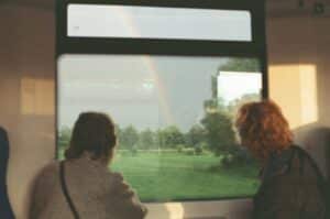 Two people, one with dark hair and the other with red, stare intently out of a train window, looking at a field surrounded by trees. A rainbow paints the gray sky.