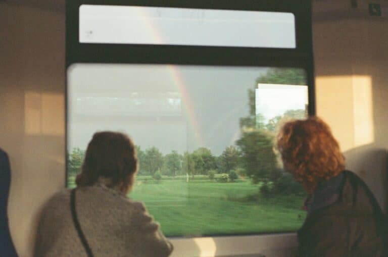 Two people, one with dark hair and the other with red, stare intently out of a train window, looking at a field surrounded by trees. A rainbow paints the gray sky.