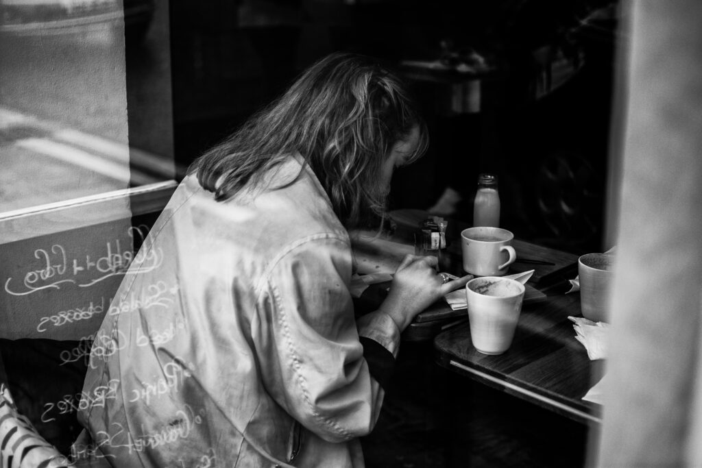 A grayscale photograph of a young woman, with frizzy hair, sitting in a coffee shop, poring over her writing. There are multiple empty coffee cups on the table beside her; she has been there a while.