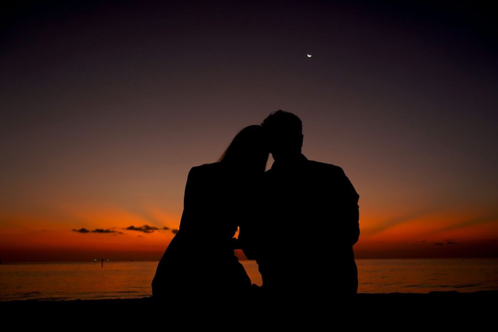 Image of a man and a woman sitting facing the sun setting over the ocean. They are sitting close and leaning against one another.