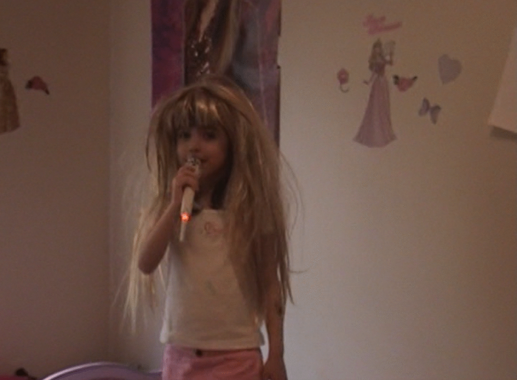 A young girl wears a blonde wig and sings into a microphone. Her room is themed after Hannah Montana.