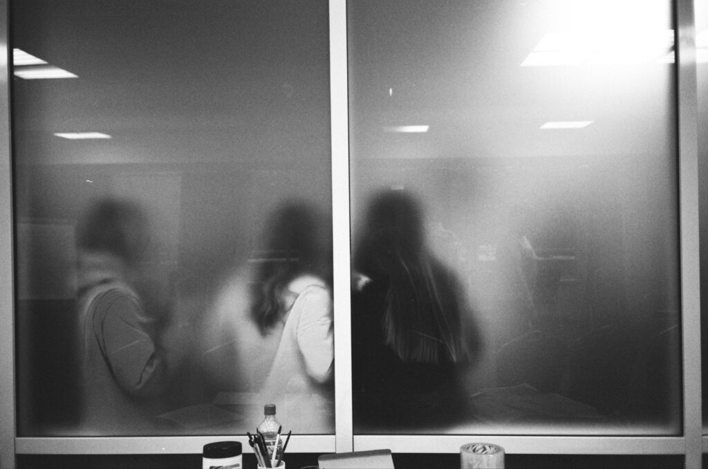 Multiple individuals, behind frosted glass, stand together chatting. They are on the other side of the pane from an office desk with pencils, paper, and other tools sitting on it.