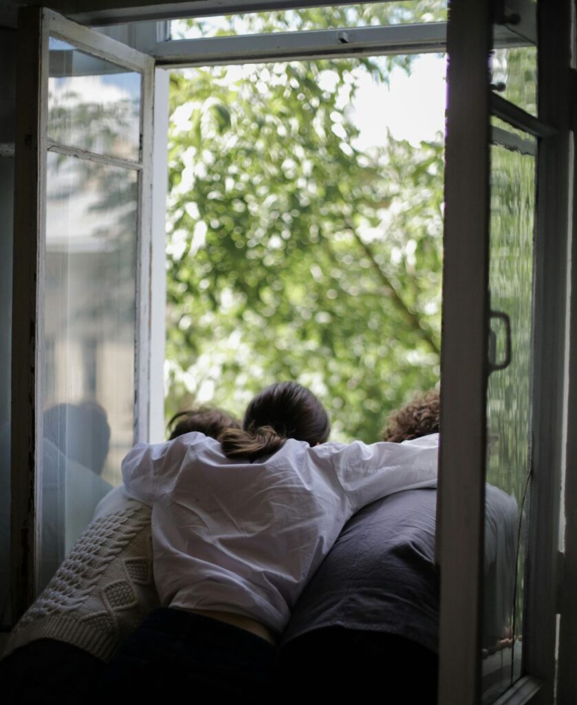 A group of friends, embracing one another, stare out a window together at green trees. They are thinking of other friends, who live far and wide, across the world.
