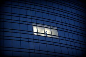 An individual stares out of the window in an office highrise. The light of the office softens their outline and loneliness against the dark backdrop.