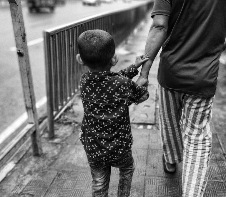 A grayscale image of a young boy, maybe five years old, grasping his parent’s arm firmly as they walk away on the sidewalk.