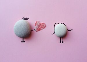A pebble with cartoon arms holding out a heart to another pebble cartoon against a pink background.